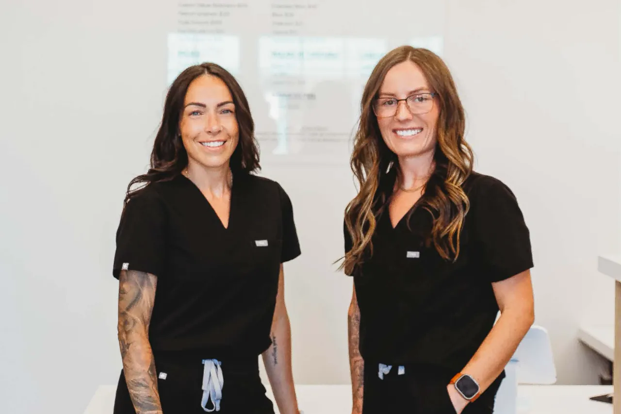 Clare Bourdon and Brooke Conrady, FNP-C, standing together at the Pomalo Wellness front desk, the service menu on the wall behind them.